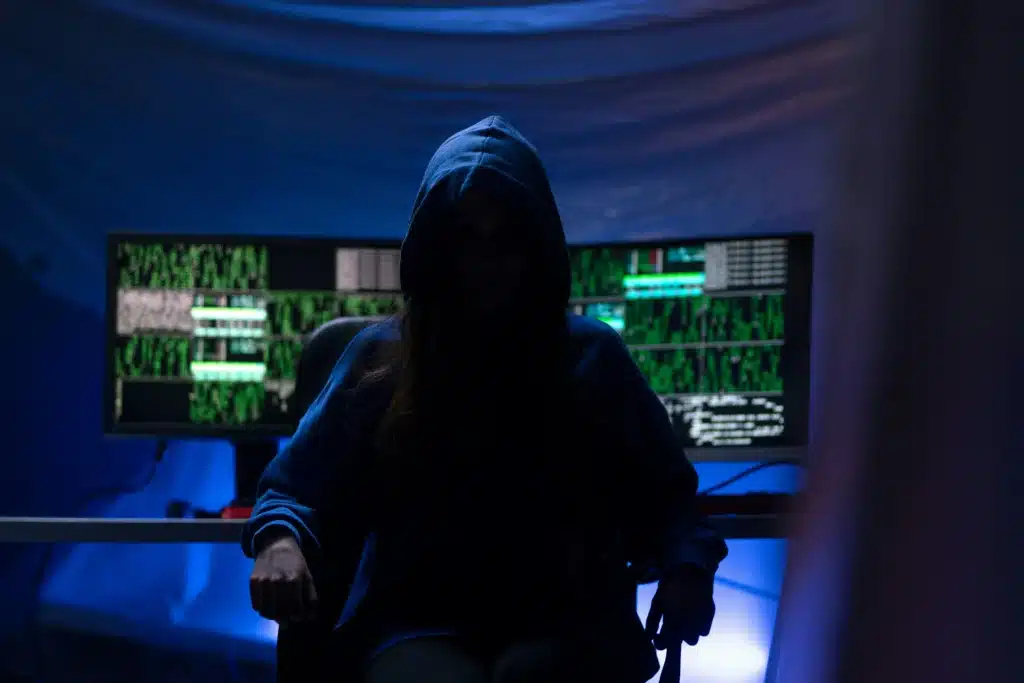 Anonymous man in blue hoodie sitting in a computer chair with green code on dual monitors at the desk behind him
