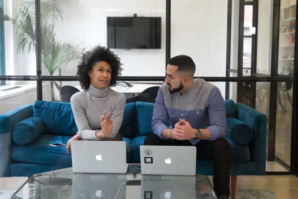 Photo of Mac IT services in an office with a man and woman talking on blue sofa in front of two Macbooks