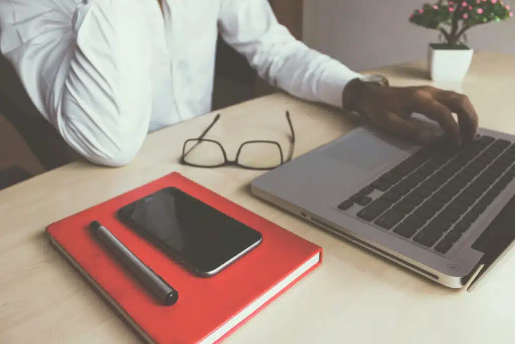 Man in white dress shirt typing on Macbook at desk next to notebook and iPhone