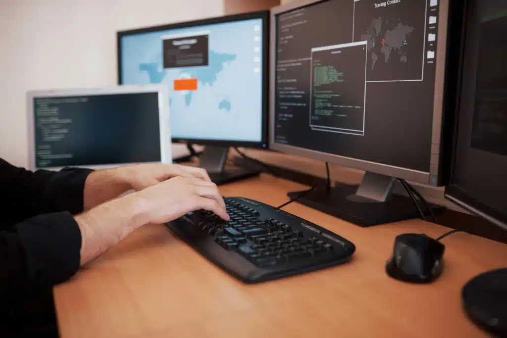 Man's hands typing on black keyboard at desk with dual monitors and laptop showing code