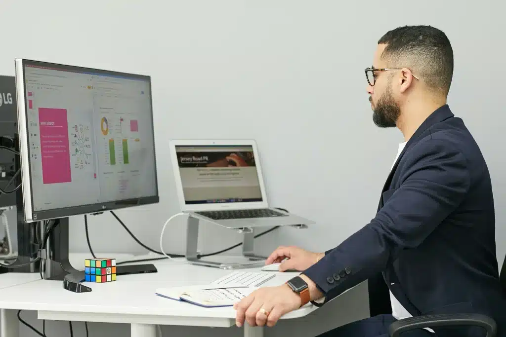 Photo of a man working at a desk on a monitor and laptop with data on the screen
