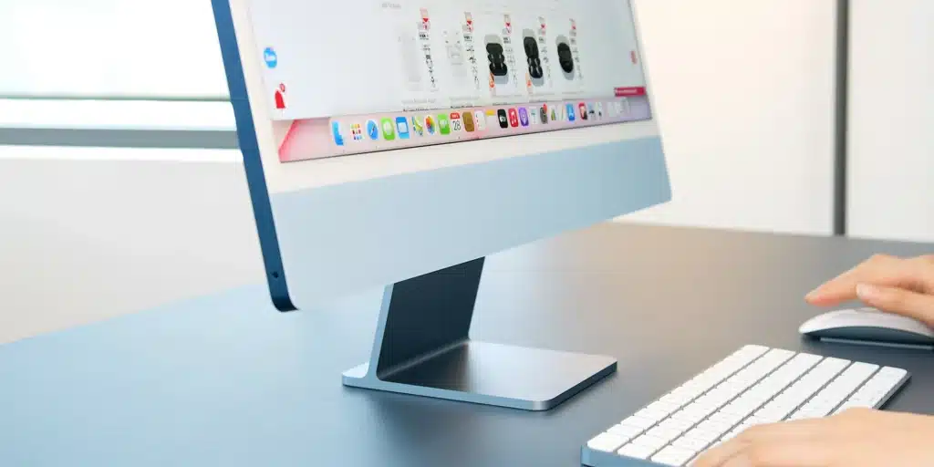Side view of iMac with apps on screen and hands typing and using mouse at grey desk