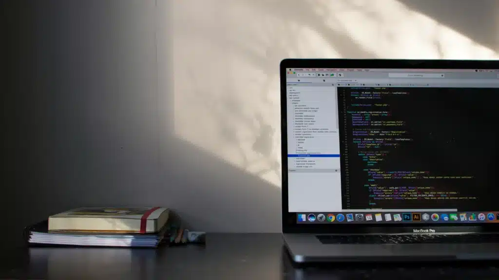 Space grey MacBook Pro open with code on screen next to stack of notebooks with pencils