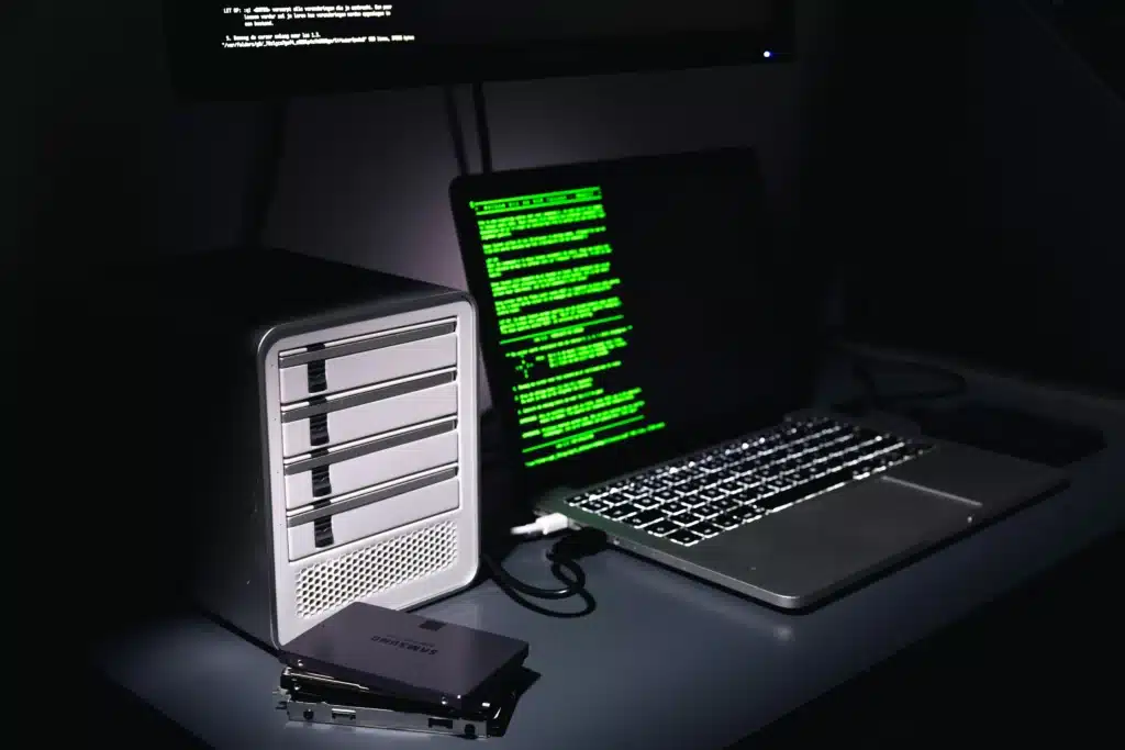 Space grey macbook pro with green code on screen, plugged into a database and storage device at a desk in a dark room