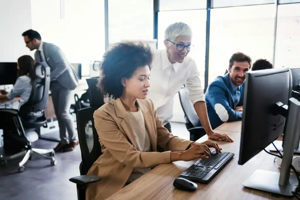 Woman typing on computer while another woman watches behind smiling in office setting with other employees dressed professionally